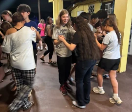 A group of teenagers dance in pairs on a covered outdoor patio at night, some smiling and holding hands, while others stand or walk in the background.