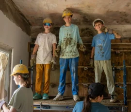 Three young people in hard hats and work clothes stand on a platform indoors, covered in dirt, while others work nearby, suggesting a group construction or renovation project.