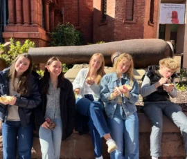 Five people sit on a stone ledge outdoors, eating bowls of food and smiling; a large sculpture and brick building are in the background.