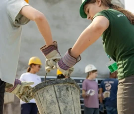 Two people wearing gloves and hard hats lift a large bucket together, with a group of others in construction gear standing in the background.