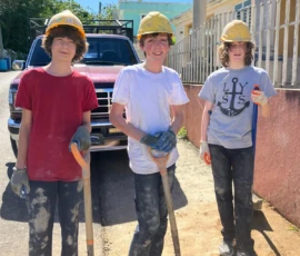 Three young people wearing hard hats and gloves stand outdoors holding shovels, with a truck and building in the background.