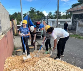 Four people wearing helmets shovel gravel from a pile on a street into buckets, with a truck and buildings visible in the background.