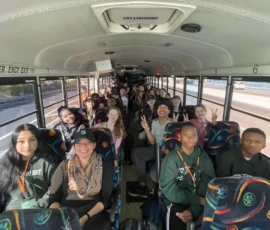 A group of students sits on a school bus, facing forward and smiling, with some making peace signs. The bus is on the road during daylight.