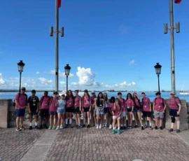 A group of people wearing matching pink shirts stands in a line on a brick walkway next to a waterfront, with lamp posts and blue sky in the background.