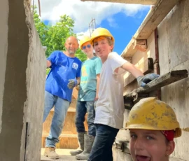 Four people wearing construction helmets and gloves stand on a building site, posing and smiling for the camera, with concrete walls and a bright blue sky in the background.