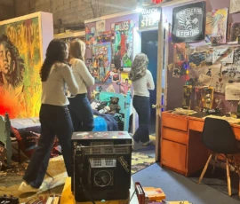 Three people walk through a cluttered, retro-style bedroom filled with posters, artwork, and various objects. Bright light shines from an open door.