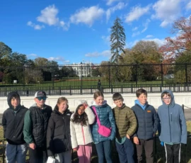 A group of eight children stand in a row outside, posing in front of the White House on a sunny day.