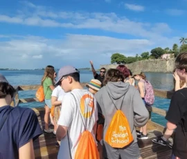 A group of students with orange drawstring bags labeled "The Saklan School" stand on a wooden dock by the water, facing an instructor, with trees and a stone wall in the background.