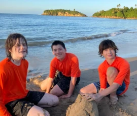 Three boys in orange shirts sit on a sandy beach building a sand structure, with the ocean and a green island visible in the background.
