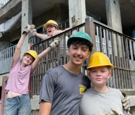 Four young people wearing construction helmets and work clothes stand in front of a partially built concrete structure, smiling and posing for the camera.