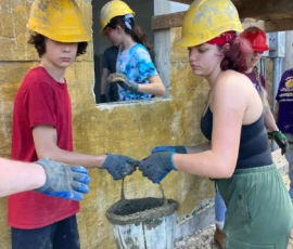 Two people in hard hats and gloves pass a bucket of cement at a construction site, with others working in the background.