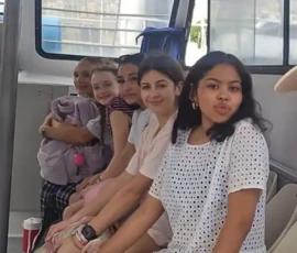 Five young women sit in a row on a boat, facing the camera. Some are smiling, and one is making a playful face. They appear relaxed and casual.