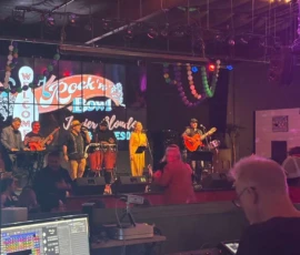 A live band performs on stage under colorful lights while a person operates a soundboard in the foreground at an indoor venue.