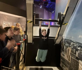 A child hangs from a horizontal bar while other children watch in a science museum exhibit with a cityscape backdrop.