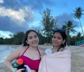 Two young women stand on a sandy beach; one wears a pink swimsuit holding items, while the other is wrapped in a light towel. Palm trees and clouds are visible in the background.
