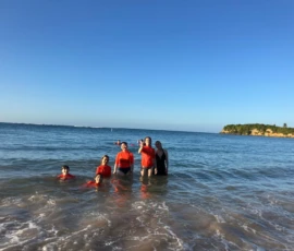 Six people stand and swim in shallow ocean water near the shore, with an island and clear blue sky visible in the background.