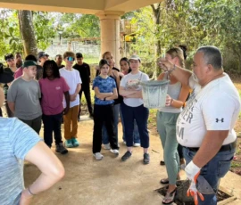 A group of people stands outside under a covered area, listening to a man holding up a bucket and wearing work gloves.