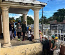 A group of people wearing hard hats and gloves gather outside a house with columns and a yellow wall, preparing for work or a project.