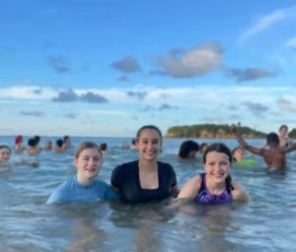 Three girls stand in the ocean water smiling at the camera, with several other people swimming and playing in the background under a blue sky.