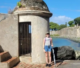 A person stands in front of a historic stone guardhouse by the water on a sunny day, holding a striped umbrella.