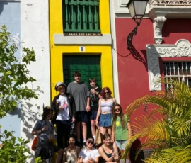 A group of people pose for a photo on steps in front of a colorful building with a green door framed in yellow and the number 1 above it.