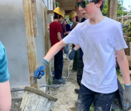 A boy in a red hard hat and sunglasses carries a bucket of cement at a construction site, with other people working in the background.