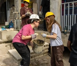 Several people wearing hard hats and gloves work together carrying cinder blocks and a bucket at a construction site.