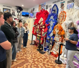 A group of people listen to a guide in a museum, standing near elaborate Mardi Gras Indian costumes displayed on mannequins against walls covered with photos and memorabilia.