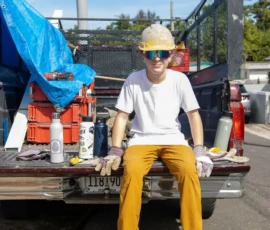 A boy wearing a hard hat, safety glasses, gloves, and casual clothes sits on the open tailgate of a pickup truck with tools and supplies in the truck bed.