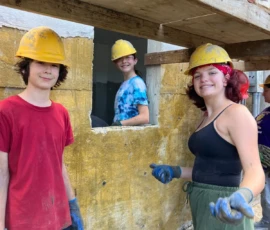 Three people wearing yellow construction helmets and gloves stand by a window opening in a building under construction, with one person inside smiling through the window.