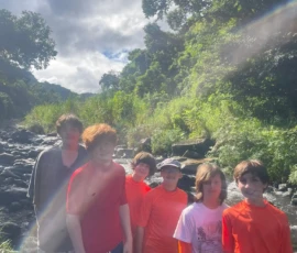 Six teens stand by a rocky creek in bright sunlight, surrounded by greenery, some wearing orange long-sleeve shirts.