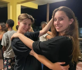 Two young people smile and pose as they dance together indoors at night, with other people socializing in the background.