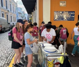 A group of people stand in line at a street cart where a vendor serves food on a cobblestone street in front of a yellow building with a "Lord Jim Leather" sign.