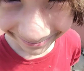 A young person with shaggy brown hair, wearing a red shirt, looks up at the camera and smiles outdoors in bright sunlight.
