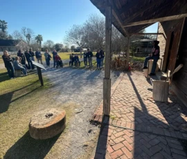 A group of people listen to a person speaking on the porch of a rustic wooden building on a sunny day, with open grassy areas and trees in the background.