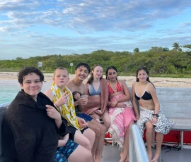 Six teenagers wrapped in towels sit together on a boat, with a sandy beach and green trees in the background under a partly cloudy sky.