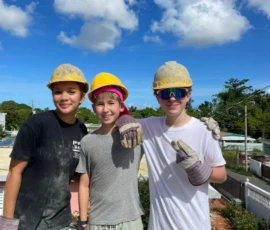 Three young people wearing yellow construction helmets, gloves, and casual clothes stand outdoors on a building site under a blue sky with clouds.