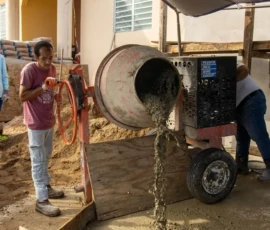 A person operates a cement mixer, pouring wet concrete onto the ground at a construction site next to a building.