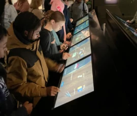 People interact with large touchscreen exhibits in a dimly lit museum, observing and engaging with digital displays.