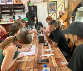 A group of people sit around a wooden table in a busy café, engaged in conversation and writing on paper, with drinks and menus visible.