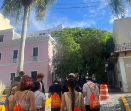 A group of students with orange drawstring bags walk together on a cobblestone street lined with palm trees and colorful buildings under a blue sky.