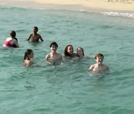A group of people swim and stand together in shallow ocean water near a sandy beach with greenery in the background.