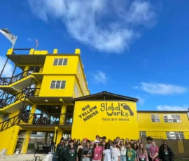 A large group of people stands in front of a bright yellow building labeled "Big Yellow House, Global Works, Travel With Purpose" under a blue sky with scattered clouds.