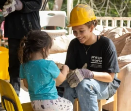 An adult wearing a yellow hard hat and gloves sits on a chair, smiling at a young child sitting in a yellow chair. Another person stands nearby, also wearing gloves.
