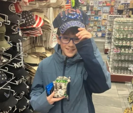 A child wearing glasses and a blue jacket tries on a sequined USA cap in a souvenir shop, holding small souvenir items in hand, surrounded by hats and other merchandise.