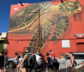 A large sea turtle mural is painted on a red building wall above parked cars while people walk by on the sidewalk below.