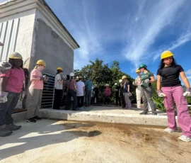 A group of people wearing hard hats and work gloves stand on a rooftop under a blue sky, appearing to participate in a construction or volunteer project.