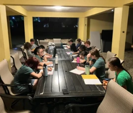 A group of people sit around a large outdoor table at night, writing or reading from notebooks under bright overhead lights.