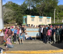 A large group of people pose together outdoors in front of a sign reading "Vega Alta, Villa del Río," with trees and a building in the background.