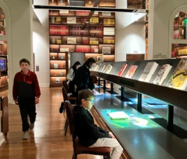 Visitors, including children, view illuminated display tables and books in a modern library room with tall, book-lined walls and a wooden floor.